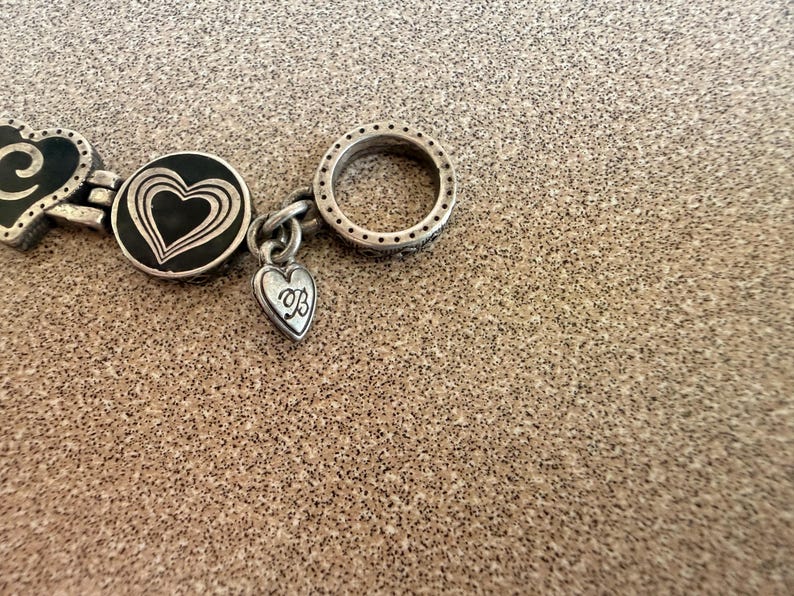 Puede incluir: Primer plano de una pulsera plateada con dijes en forma de coraz&oacute;n. La pulsera presenta un eslab&oacute;n circular con peque&ntilde;os detalles de puntos y un dije en forma de coraz&oacute;n con una inicial. La pulsera est&aacute; sobre una superficie texturizada.