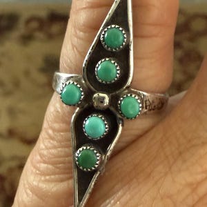 Puede incluir: Un anillo plateado con un diseño central alargado y cinco piedras de color turquesa. El anillo tiene un fondo oscuro oxidado, y las piedras están engastadas en un patrón decorativo. Una pieza de joyería única.