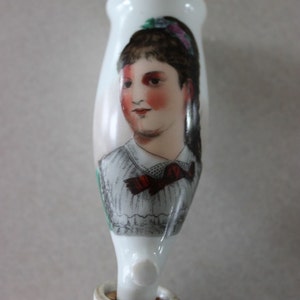 May include: White porcelain figurine of a woman with a red bow and a floral headband. The figurine is painted with a detailed portrait of the woman's face.