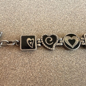 Puede incluir: Pulsera plateada con abalorios en forma de coraz&oacute;n y cuadrados con detalles de esmalte negro. La pulsera tiene un cierre de palanca y una serie de dise&ntilde;os de coraz&oacute;n, incluyendo un cuadrado con un coraz&oacute;n, un coraz&oacute;n con un remolino y un coraz&oacute;n circular.