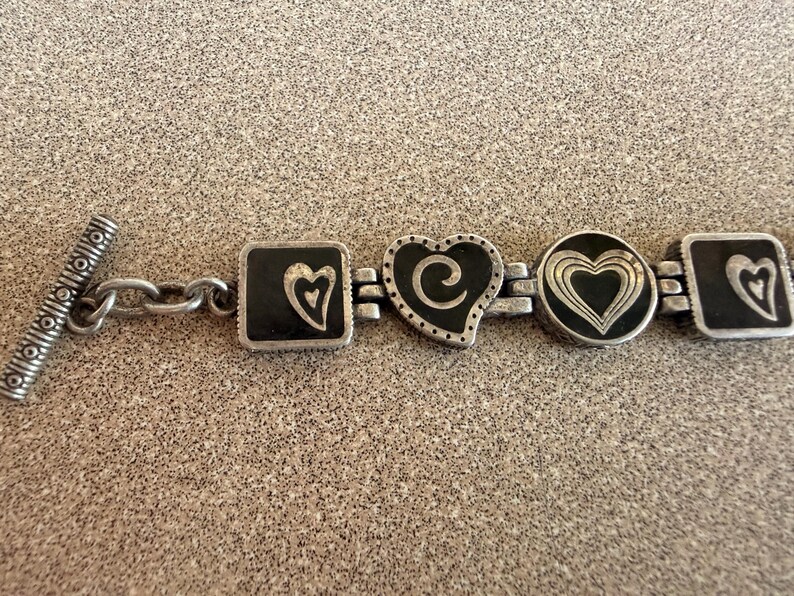 Puede incluir: Pulsera plateada con abalorios en forma de coraz&oacute;n y cuadrados con detalles de esmalte negro. La pulsera tiene un cierre de palanca y una serie de dise&ntilde;os de coraz&oacute;n, incluyendo un cuadrado con un coraz&oacute;n, un coraz&oacute;n con un remolino y un coraz&oacute;n circular.