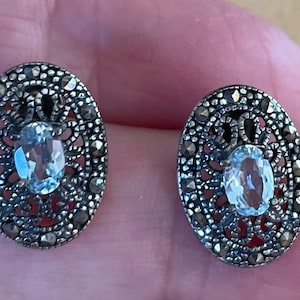 May include: A pair of silver earrings featuring a delicate filigree design and a light blue oval gemstone in the centre.
