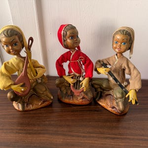 Pode incluir: Três figuras vintage de músicos. Cada figura está ajoelhada sobre uma base, vestindo uma roupa de cor diferente e segurando um instrumento musical. As figuras têm rostos pintados e são feitas de cerâmica.