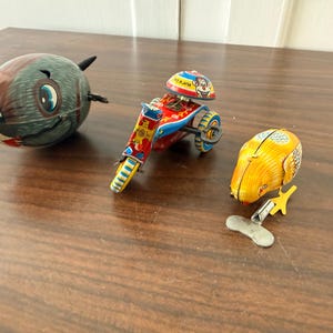 May include: A collection of three vintage tin toys including a grey, oval-shaped fish with blue eyes, a red, blue, and yellow motorcycle with a clown figure, and a yellow bird with a metal wind-up key attached to its base for mechanical movement.