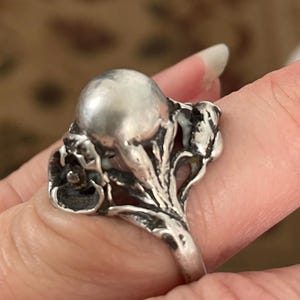 Könnte beinhalten: Ein silberner Ring mit einem großen, runden Blumenmuster. Der Ring hat eine große, runde, silberne Kugel in der Mitte, umgeben von zarten, silbernen Blumendetails.