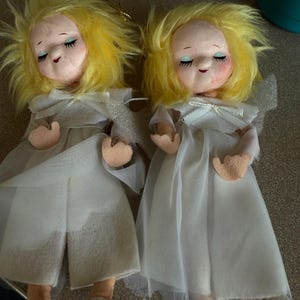 May include: Two vintage angel dolls with yellow yarn hair, closed eyes, and rosy cheeks. They wear white dresses with sheer overlay and small bows. The dolls have fabric hands and feet.
