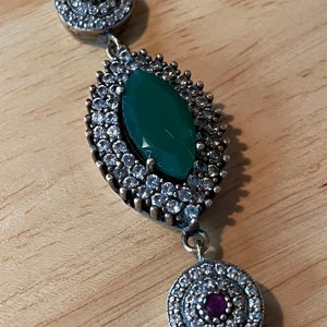 Peut inclure: Un bracelet en argent avec une grande pierre précieuse verte entourée de petites pierres claires. Le bracelet a également deux petits pendentifs ronds avec des pierres claires et une seule pierre rouge au centre.