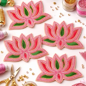 May include: Four pink lotus flower appliques with red and green accents are displayed on a white surface. Gold scissors, beads, and spools of thread surround the appliques, suggesting a crafting theme. The appliques are embellished with beads.