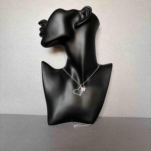 May include: A silver necklace with a heart pendant and a butterfly accent. The pendant is displayed on a black mannequin bust against a gray background. The necklace chain is delicate and the heart is open. The butterfly is on the right side of the heart.