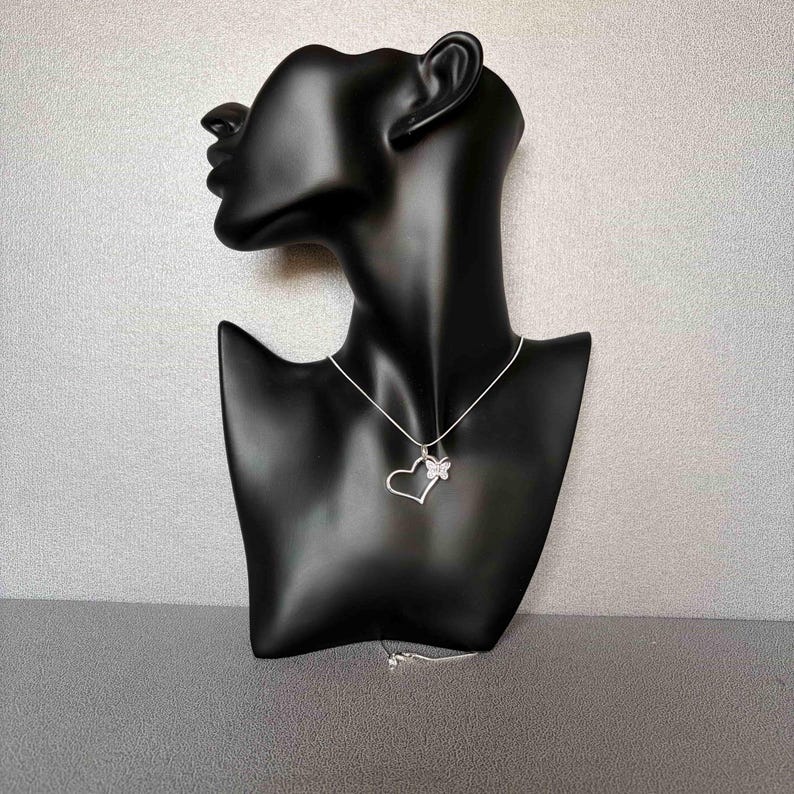 May include: A silver necklace with a heart pendant and a butterfly accent. The pendant is displayed on a black mannequin bust against a gray background. The necklace chain is delicate and the heart is open. The butterfly is on the right side of the heart.