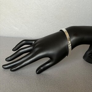 May include: A silver-colored bangle bracelet with a textured surface. The bracelet is displayed on a black hand mannequin against a gray background. The bracelet's design features small, repeating patterns.
