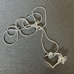 May include: A silver-colored necklace with a heart-shaped pendant and a butterfly accent. The pendant is open in the center and has a small butterfly attached to the right side. The chain is a thin, silver snake chain.