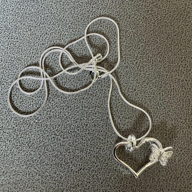 May include: A silver-colored necklace with a heart-shaped pendant and a butterfly accent. The pendant is open in the center and has a small butterfly attached to the right side. The chain is a thin, silver snake chain.