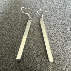 May include: A pair of silver-toned drop earrings featuring long, slender, rectangular bar pendants. Each earring is attached to a curved wire hook, creating a simple, elongated vertical design with a smooth, reflective metallic finish.