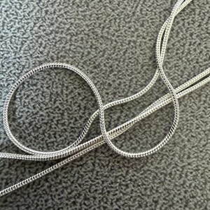 May include: Close-up of a silver-colored chain necklace with a snake chain design. The chain is coiled in a circular pattern and lies on a gray textured surface. The necklace is made of a shiny, reflective metal.