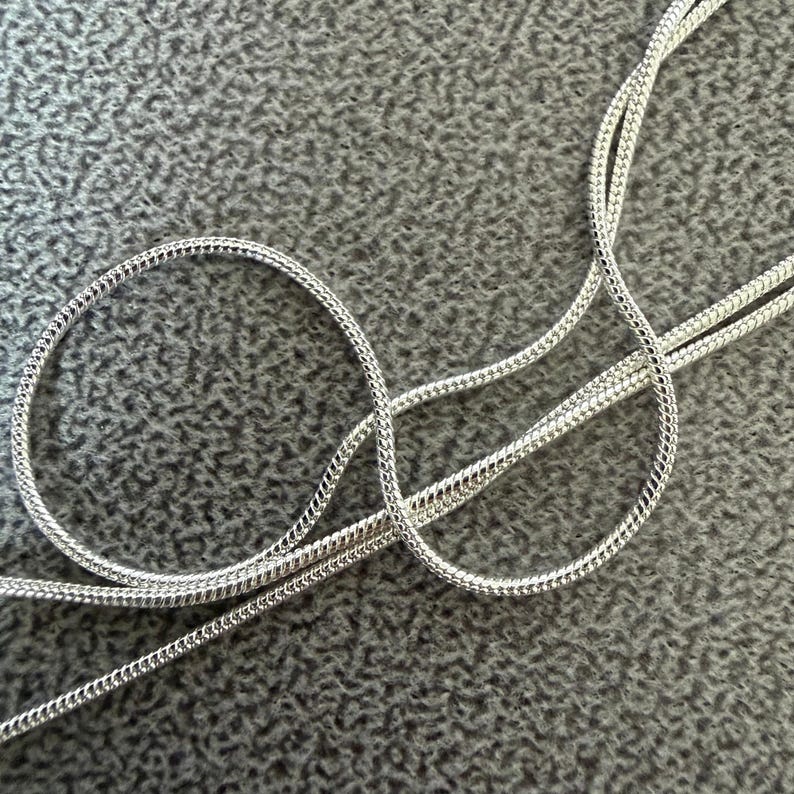 May include: Close-up of a silver-colored chain necklace with a snake chain design. The chain is coiled in a circular pattern and lies on a gray textured surface. The necklace is made of a shiny, reflective metal.
