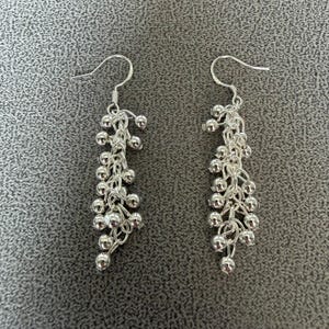 May include: A pair of silver-toned dangle earrings featuring a cluster of small, round metallic beads suspended from delicate silver chains. Each earring is attached to a curved ear wire hook, creating a cascading, textured, and reflective design.