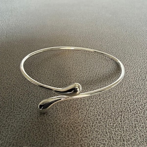 May include: A silver-colored cuff bracelet with a minimalist design. The bracelet features a slender, open band that curves around the wrist, with teardrop-shaped accents at each end. The smooth, polished metal reflects light.