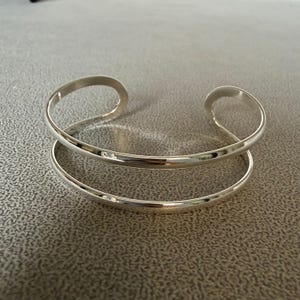 May include: A silver-colored cuff bracelet with a double-band design. The bracelet has a smooth, polished finish and a curved opening. The bands are approximately 0.5 inches wide. The bracelet is displayed on a neutral-toned surface.