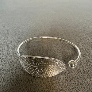 May include: A silver-colored bangle bracelet with a leaf design. The leaf is detailed with intricate veining and is attached to the bracelet's curved band. The bracelet has a small, round ball at the opening.