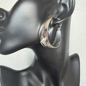 May include: A large, polished silver-tone hoop earring with a wide, flat band design. The earring features a smooth, reflective metallic surface and a secure hinged post closure, displayed on a black mannequin ear and neck form.