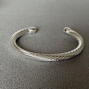 May include: A silver-toned cuff bracelet featuring a continuous twisted cable pattern throughout the band. The bracelet has a circular opening and terminates at both ends with smooth, rounded, polished silver-toned metal caps.