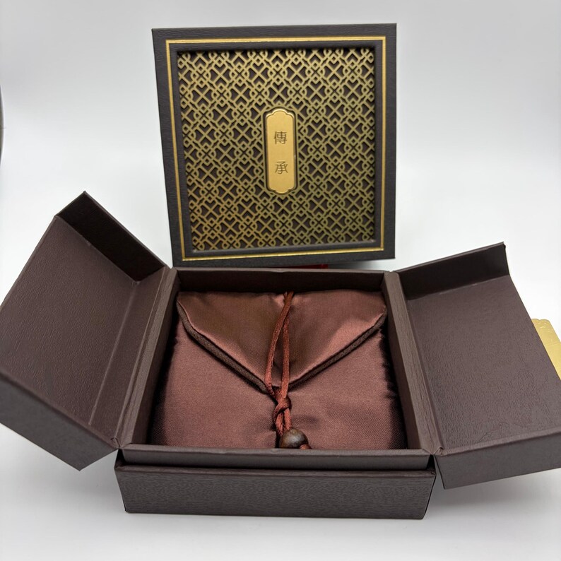 May include: An open, brown gift box with a satin pouch inside. The box has a decorative gold and black patterned lid with a gold plaque that has Chinese characters. The pouch is a deep red-brown color and is tied with a matching cord.