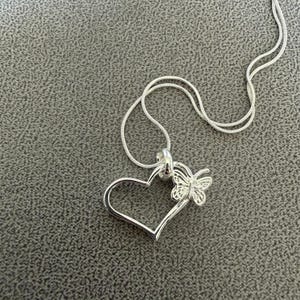 May include: A silver-colored necklace with a heart-shaped pendant and a butterfly accent. The pendant is open, with a delicate butterfly design attached to the side of the heart. The chain is thin and silver.