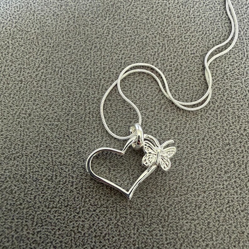 May include: A silver-colored necklace with a heart-shaped pendant and a butterfly accent. The pendant is open, with a delicate butterfly design attached to the side of the heart. The chain is thin and silver.