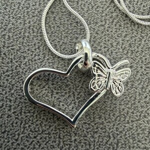 May include: A silver-toned pendant necklace featuring a heart-shaped design with a butterfly accent. The butterfly is positioned on the right side of the heart. The necklace chain is also silver-toned and appears to be a snake chain.