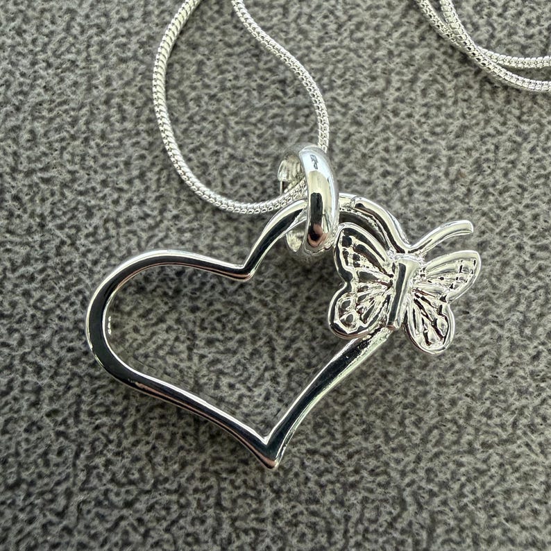 May include: A silver-toned pendant necklace featuring a heart-shaped design with a butterfly accent. The butterfly is positioned on the right side of the heart. The necklace chain is also silver-toned and appears to be a snake chain.