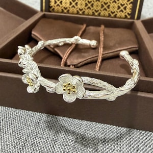 May include: Silver-toned floral cuff bracelet. The bracelet features a branch-like design with delicate flowers. The flowers have gold-colored centers. The bracelet is displayed in a brown jewelry box.