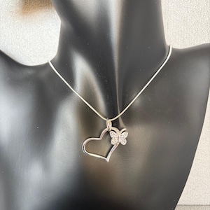 May include: A silver-toned necklace featuring a heart-shaped pendant with a butterfly accent. The heart is open, and the butterfly is embellished with small, sparkling stones. The necklace is displayed on a black mannequin.
