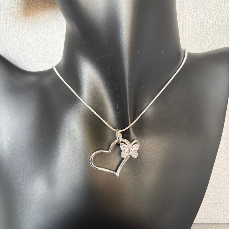May include: A silver-toned necklace featuring a heart-shaped pendant with a butterfly accent. The heart is open, and the butterfly is embellished with small, sparkling stones. The necklace is displayed on a black mannequin.