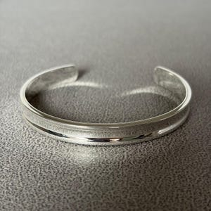 May include: A polished silver-tone cuff bracelet featuring a smooth, curved exterior surface with a subtle central horizontal groove that runs along the entire length of the band, creating a reflective and clean metallic appearance.