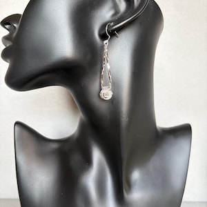 May include: A silver-toned drop earring featuring a slender, twisted open teardrop frame that suspends a small, round, pavé-set crystal charm at the base. The earring is attached to a simple hook-style finding for pierced ears.