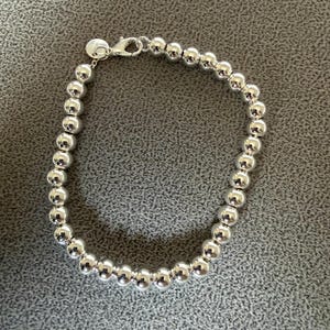 May include: A silver-toned beaded bracelet featuring a continuous strand of polished, spherical beads. The bracelet is secured with a lobster claw clasp and includes a small, circular metal tag attached near the closure mechanism.