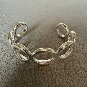 May include: A silver-colored cuff bracelet featuring a series of connected oval links. The open-ended design allows for easy wear. The bracelet has a polished, reflective surface, giving it a modern and elegant look.