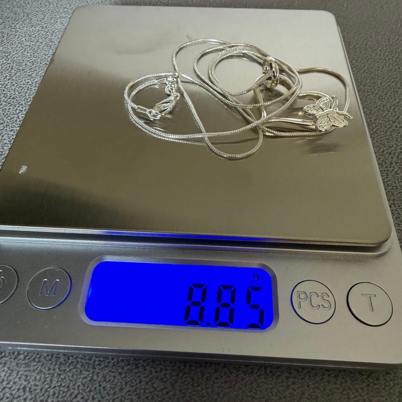 May include: A silver necklace with a butterfly pendant rests on a digital scale. The scale displays a weight of 8.85 grams. The necklace chain is a delicate snake chain style, and the butterfly pendant is detailed.