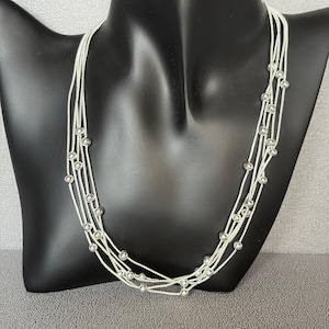 May include: A multi-strand silver necklace with small, round, silver beads evenly spaced along each strand. The necklace is displayed on a black mannequin, showcasing its layered design and delicate details. The necklace is a stylish accessory.