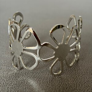 May include: A silver-toned cuff bracelet featuring a repeating openwork floral pattern. The design consists of interconnected petal shapes forming a continuous band with a polished, reflective metallic finish and an open back for adjustment.