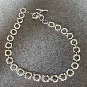 May include: A silver-toned chain necklace composed of a series of interconnected, rounded square links. The necklace features a toggle clasp mechanism consisting of a horizontal bar and a circular ring, providing a secure closure for the piece.