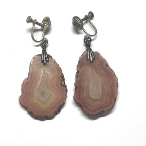 May include: A pair of dangle earrings featuring irregular, slice-cut agate stones in shades of dusty rose and cream. The stones are suspended from silver-toned metal screw-back findings, showcasing natural concentric banding patterns within each piece.