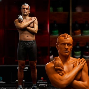 May include: A detailed sculpture of a man with a bird perched on his arm, with a matching bust. The full figure is standing on a brick and concrete base, wearing shorts and trainers. The bust is orange, and the full figure is painted with realistic skin tones.