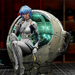 May include: A detailed figurine of a character with blue hair, wearing a white and gray suit, seated in a futuristic chair. The chair is metallic with intricate details and a glowing green interior. The figurine has a serious expression.