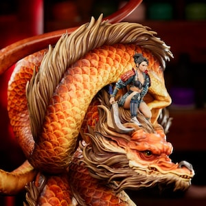 May include: A detailed figurine of a dragon with orange and gold scales, a flowing mane, and a fierce expression. A warrior figure in blue and black attire is perched atop the dragon, holding a sword. The sculpture has a dynamic pose and intricate details.