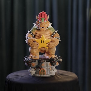 May include: A detailed figurine of Bowser, a character from the Super Mario video game series. The figure is holding a yellow star and is standing on a castle-like base. The figure is primarily orange and green with black accents.