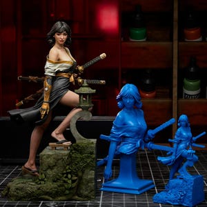 Op de afbeelding: Gedetailleerd beeldje van een vrouw in traditionele Japanse kleding, met twee zwaarden, met een miniatuur stenen lantaarn. Twee blauwe beelden van dezelfde figuur zijn ook aanwezig, met verfpotten op de achtergrond.