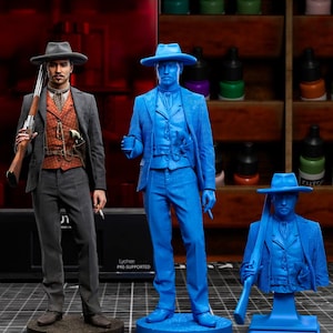 Puede incluir: La imagen muestra tres figuras: un vaquero pintado, un modelo azul y un busto azul. La figura pintada lleva un traje gris, un chaleco estampado y un sombrero, sosteniendo un rifle. Las figuras azules están en poses similares.