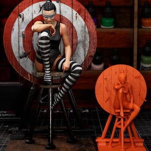 Peut inclure: Figurine détaillée d'une personne assise sur un tabouret devant une cible. La personne porte un justaucorps noir, des chaussettes hautes rayées et un bandeau. La cible est rouge et blanche. Une deuxième figurine orange est également visible.